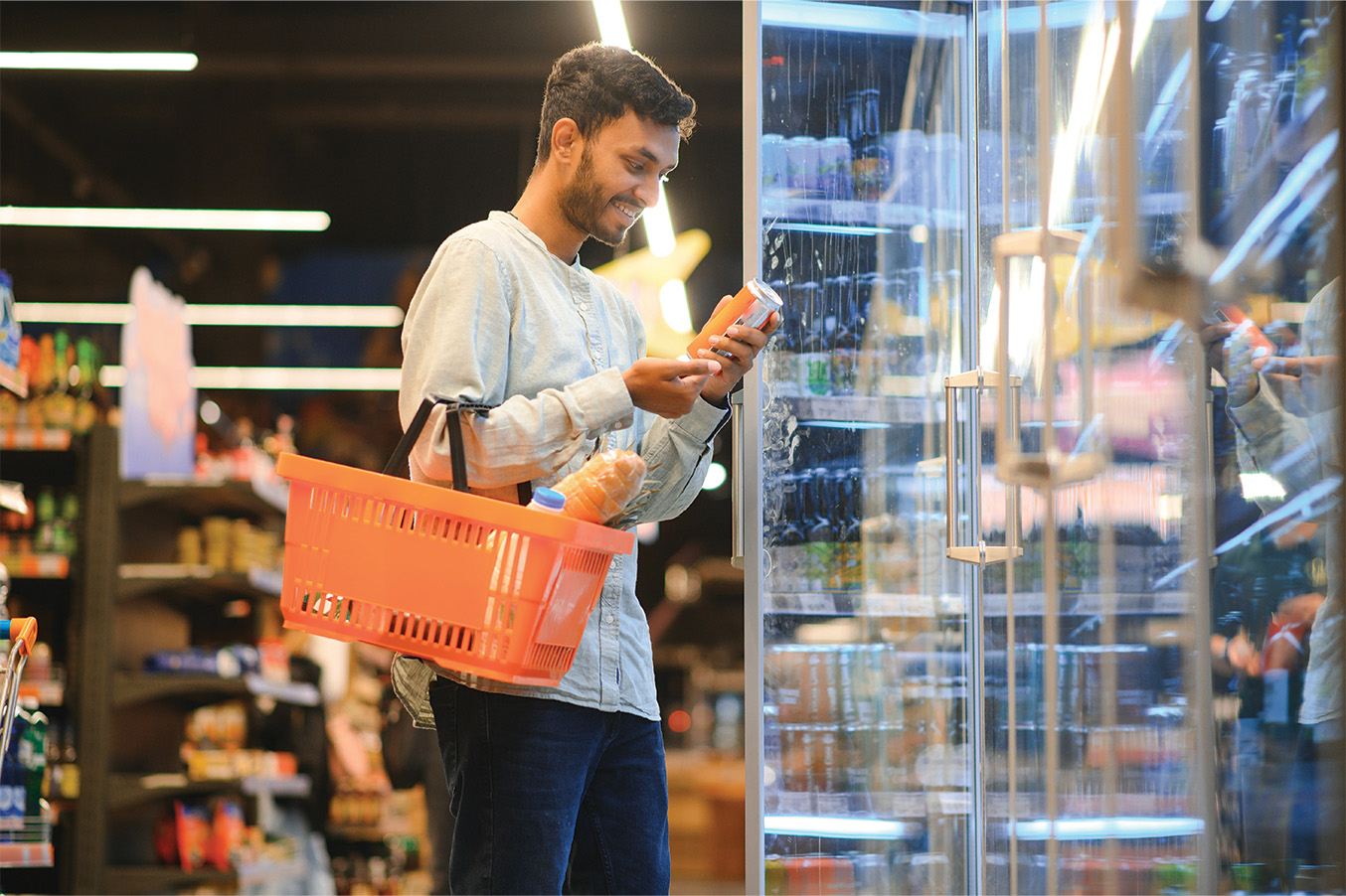 A man reading a product label at a store cooler while holding a shopping basket