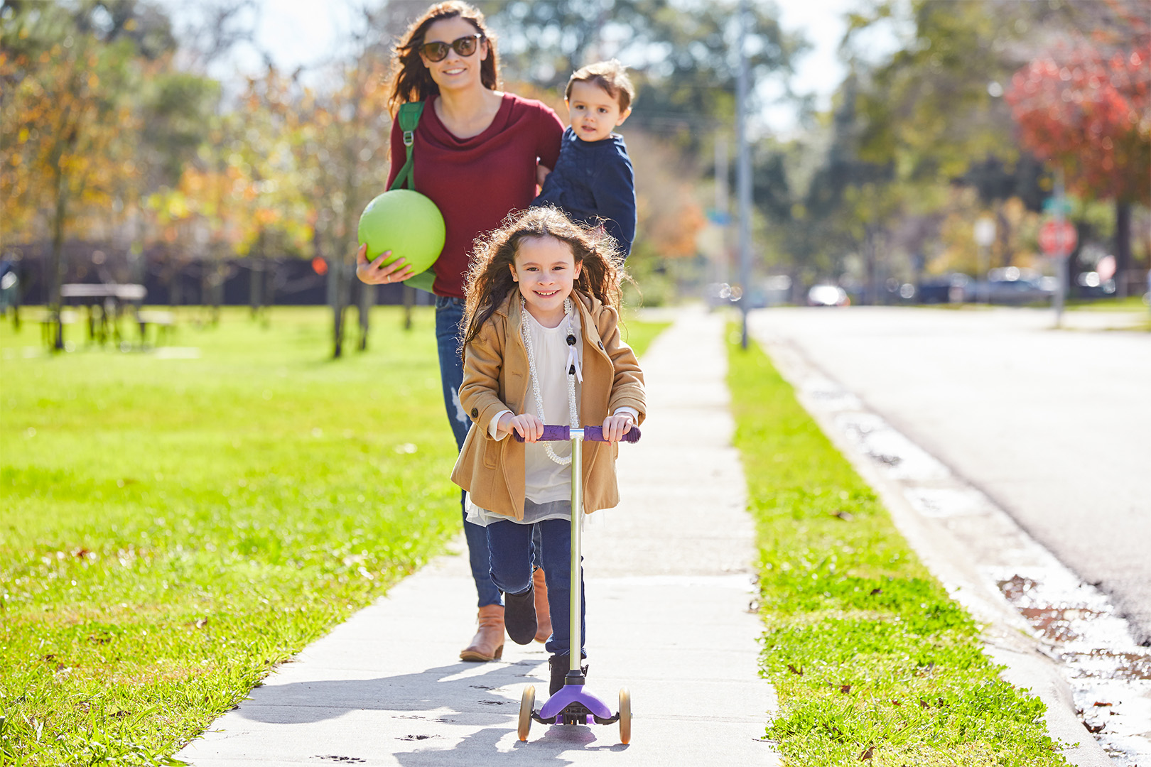 A mother and her two young children walking on a neighborhood sidewalk, one child riding a scooter